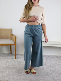 A woman wears a beige short-sleeve top, beige sandals, and the Scout Jersey Crop Flare Pant in Emerald Cove. She stands indoors on a light rug, with a wooden chair and a white table in the background.