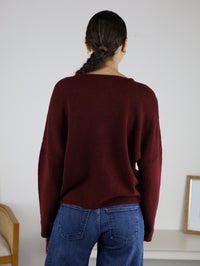 A person with dark hair in a low braid, wearing the Beth Lightweight Cardigan in maroon and blue jeans, stands facing away in a minimal room featuring a chair and a framed picture on a white shelf.