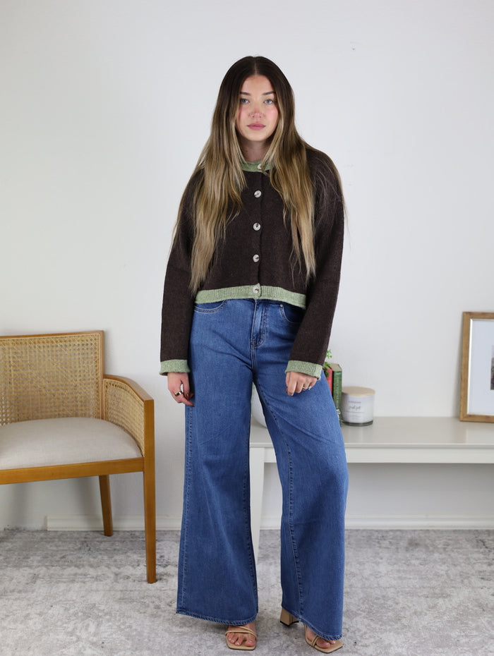 A person wears a brown, boxy fit Maya Contrast Hem Cardigan with light green trim at the hem and cuffs, paired with wide-leg blue jeans.