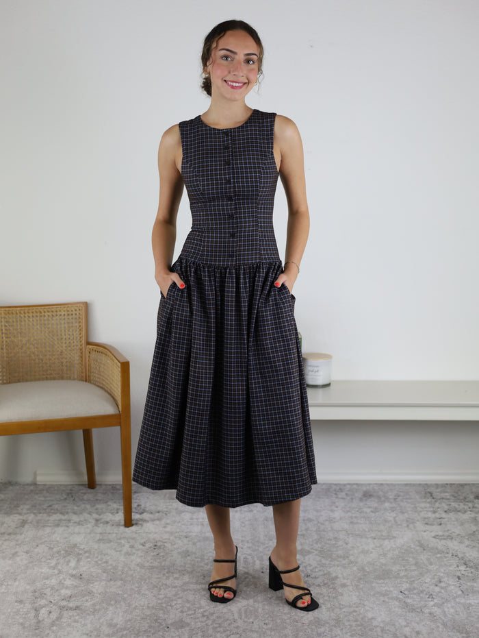 A woman stands indoors, smiling and wearing the Vivi Round Neck Button Front Midi Dress, which features a sleeveless design, dark plaid pattern, and flared skirt. She has her hands in the dress’s pockets and pairs it with black strappy heels. A bench and a chair are visible in the background.