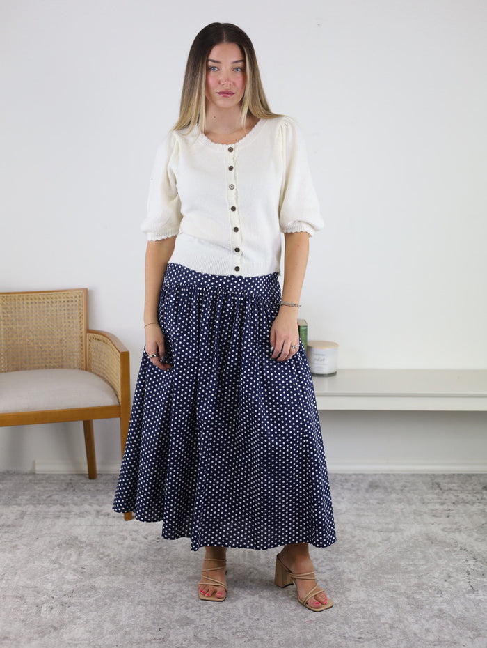 A woman stands indoors wearing the Monica Short Sleeve Cardigan—a white buttoned blouse with metallic buttons—paired with a navy blue, white polka dot maxi skirt and sandals. She is in front of a light wall, with a chair and small table in the background.