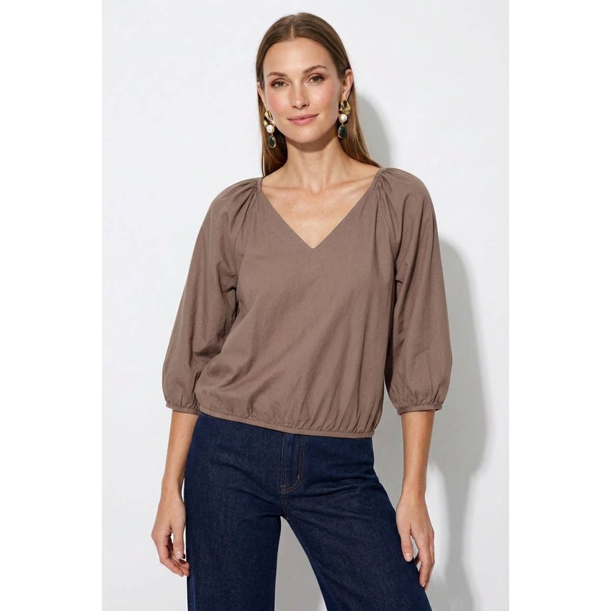 A woman wearing a Naked Zebra - Elastic Puff Sleeve V-neck Blouse: Burgundy / L, featuring three-quarter length puff sleeves and an elasticized waist, paired with dark blue jeans.