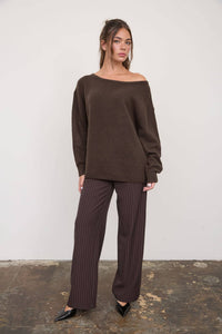 A woman stands against a plain wall wearing The Workshop Studio’s Boat Neck Oversized Knit Pullover Sweater in Red Wood, styled off the shoulder with loose brown pinstripe pants and black pointed shoes. Her hair is half-up, her expression neutral, and the floor beneath her appears worn.