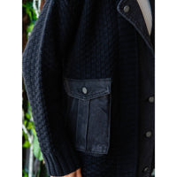 A close-up of a person wearing the Veveret - Knit Body Denim Layerd Jacket: Cream / Denim / L, featuring large front pockets and silver buttons, with their hand near the pocket and green leaves blurred in the background.