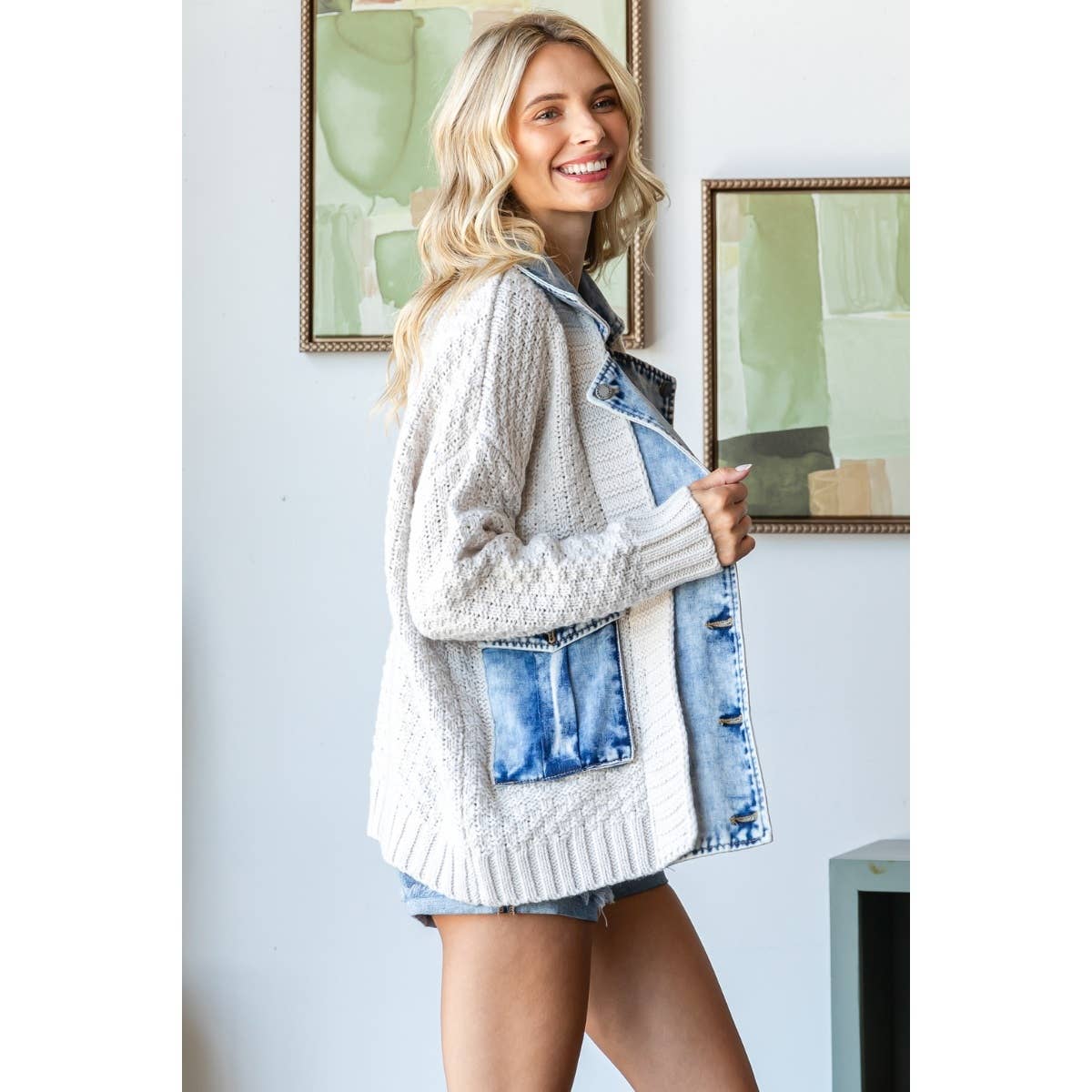 Smiling woman with long blonde hair wears a Veveret - Knit Body Denim Layerd Jacket: Cream / Denim / L and shorts, standing indoors near abstract art on a white wall.