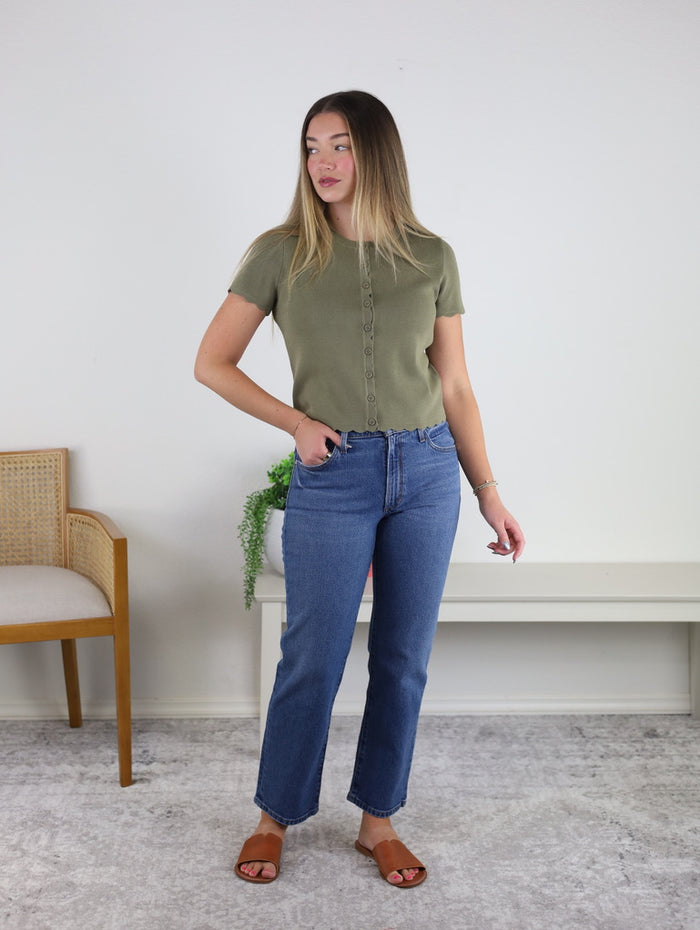 A woman stands indoors wearing the Aspen Scalloped Cardigan, styled with blue jeans and brown sandals. She looks to the side with one hand on her hip. A wooden chair and a plant on a bench are in the background.