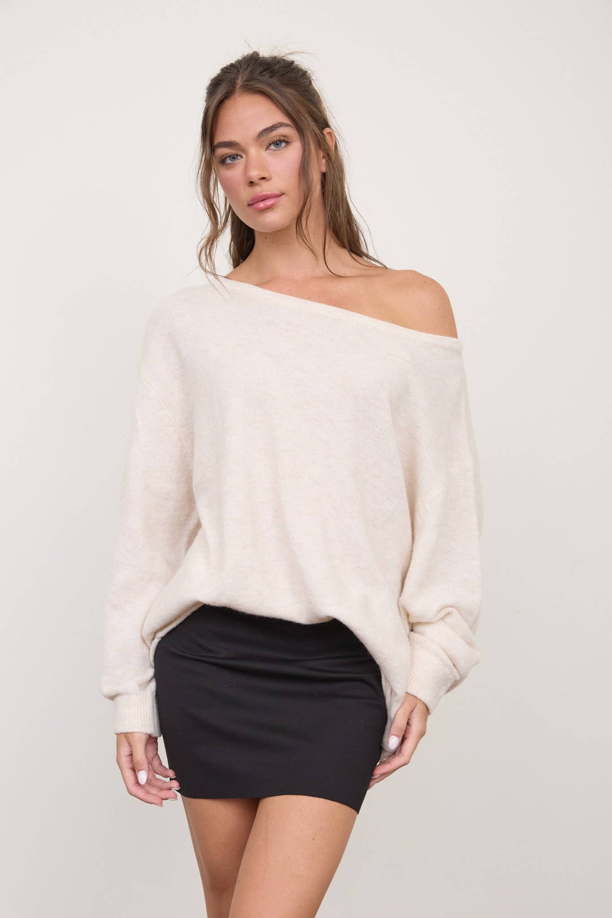 A woman stands against a plain light background, looking at the camera with a neutral expression, wearing The Workshop Studio’s Boat Neck Oversized Knit Pullover Sweater in Red Wood and a black mini skirt.