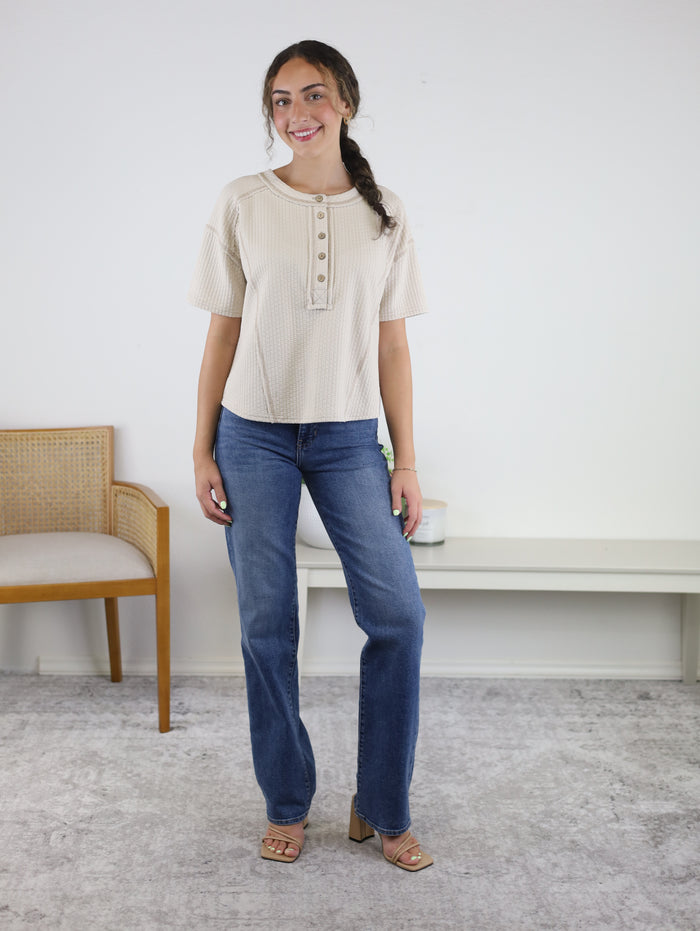 A woman with long dark hair styled in a braid stands indoors, smiling. She is wearing the Flynn Henley Knit Top in beige, which features short sleeves, paired with blue jeans and heeled sandals. A chair and a bench are visible in the background.