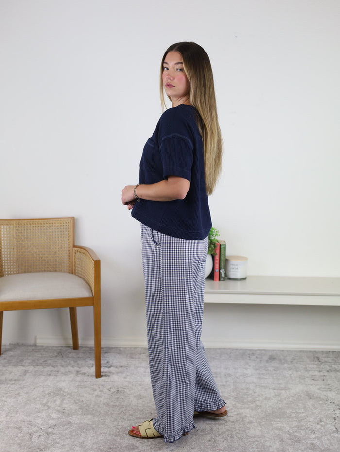 A woman with long hair stands sideways in a room, wearing a navy top, Willa Gingham Ruffle Hem Cropped Pants, and sandals. There is a wooden chair, a white shelf with books, and a plant in the background.
