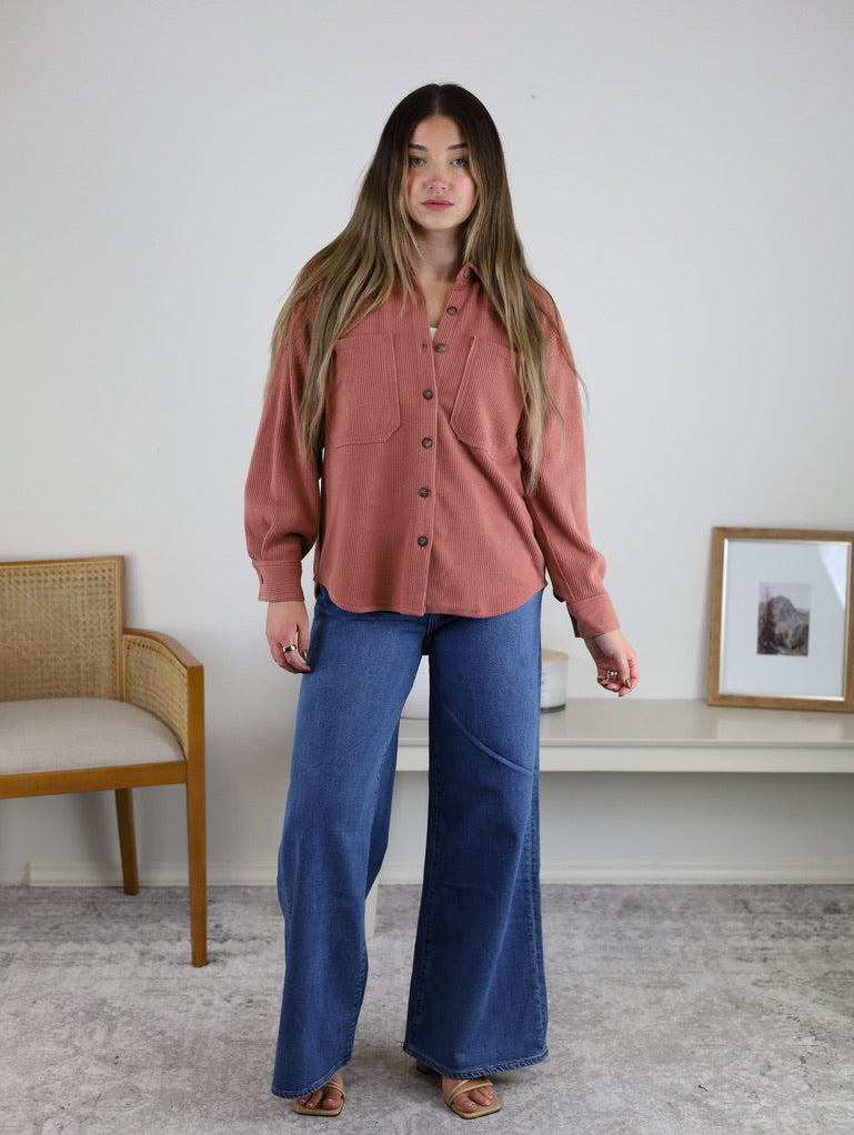 A woman with long hair wears the Larkin Rib Knit Button Up in dusty rose with wide-leg blue jeans. She stands indoors on a gray rug near a wooden chair, with framed art on a white shelf behind her.