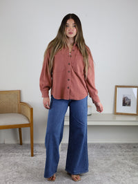 A woman with long hair wears the Larkin Rib Knit Button Up in dusty rose with wide-leg blue jeans. She stands indoors on a gray rug near a wooden chair, with framed art on a white shelf behind her.