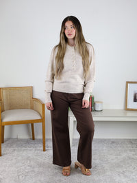 A woman with long hair wears the Theo Sweater and brown wide-leg pants, standing indoors by a chair and a white table with decor, against a plain white wall.