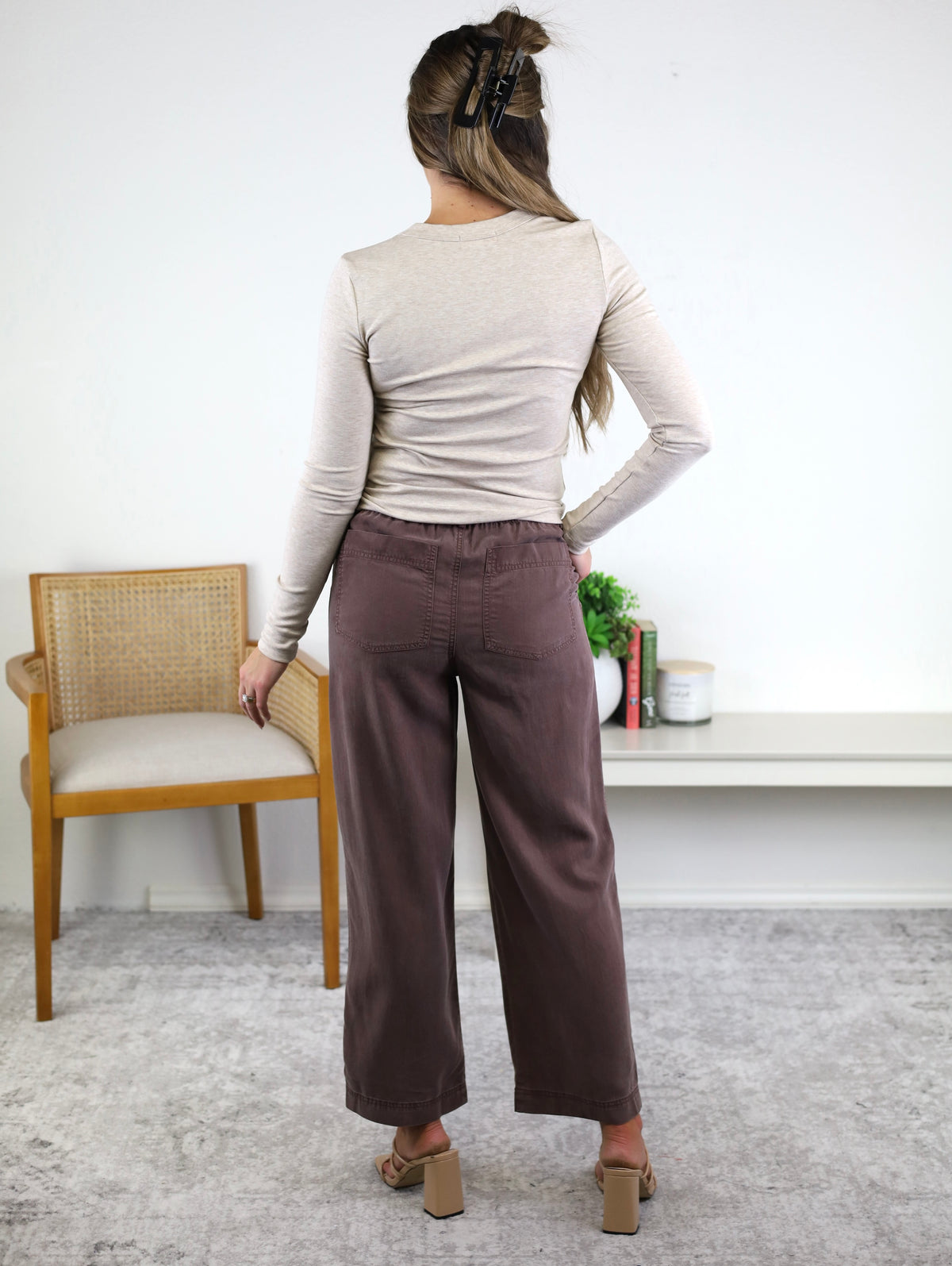 A woman stands indoors facing away, wearing a beige long-sleeve top paired with the Marbella Wide Leg Tencel Pant, featuring a drawstring elastic waist, and tan sandals. Her long hair is clipped back as she rests one hand on her hip, with a chair and shelf visible in the background.