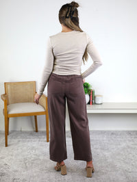A woman stands indoors facing away, wearing a beige long-sleeve top paired with the Marbella Wide Leg Tencel Pant, featuring a drawstring elastic waist, and tan sandals. Her long hair is clipped back as she rests one hand on her hip, with a chair and shelf visible in the background.