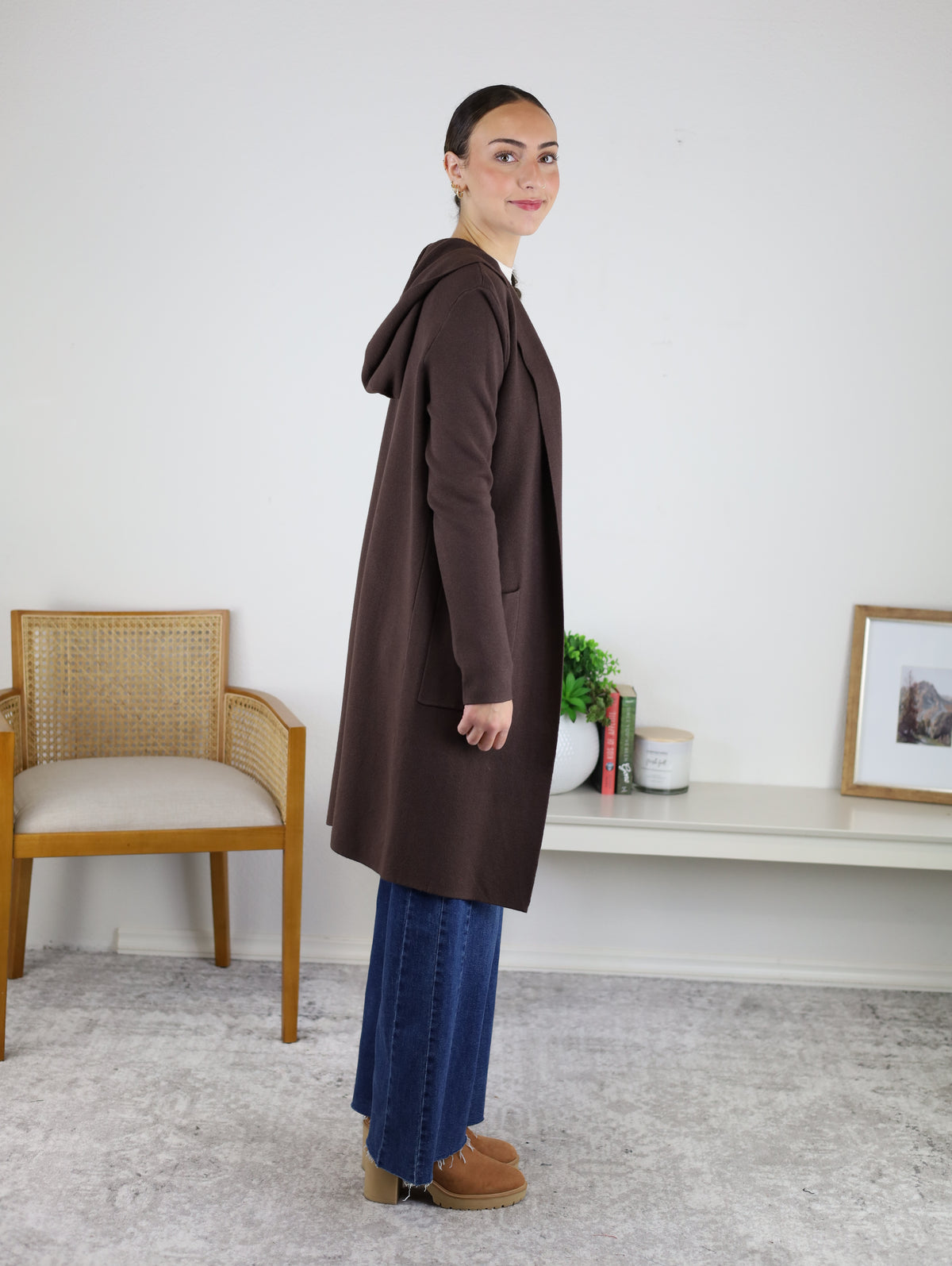 A woman stands sideways indoors, wearing the Pinch Me Long Cardigan—a cozy brown hooded layering piece—styled with blue jeans and tan shoes. Behind her are a light chair, a bookshelf, and small decorative items.
