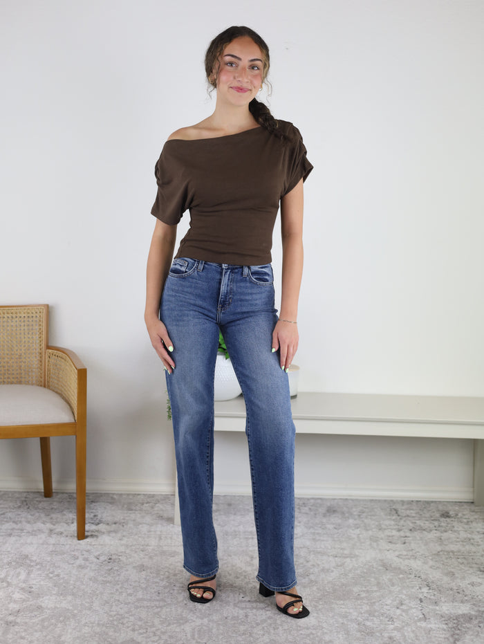 A woman stands indoors, smiling softly as she wears the Meadow One Shoulder Top in brown, paired with blue jeans and black heeled sandals. Her long, dark hair is styled in a braid. In the background, there is a beige chair and a white table.