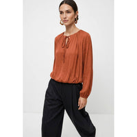 Naked Zebra - Long Sleeve Draped Bubble Hem Blouse in wine, a rust-colored, long-sleeve satin top featuring a tie-neck detail and a draped bubble hem, worn with dark trousers.