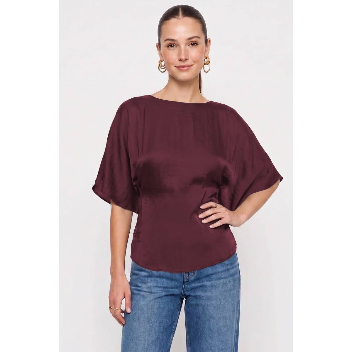 A woman wears a wine-colored, silky Naked Zebra - Back Tie Dolman Sleeve Silky Fall Blouse with a boat neck and blue jeans.
