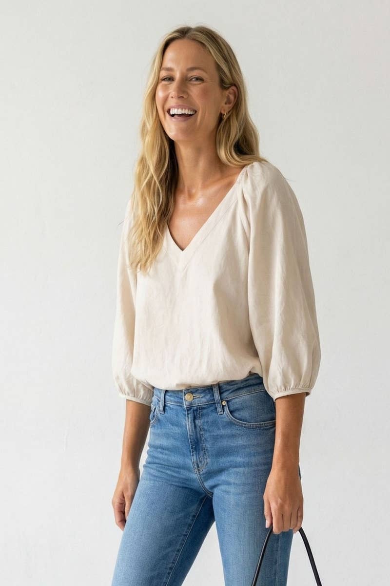 A woman wearing a Naked Zebra - Elastic Puff Sleeve V-neck Blouse in cream with puff sleeves and blue jeans.