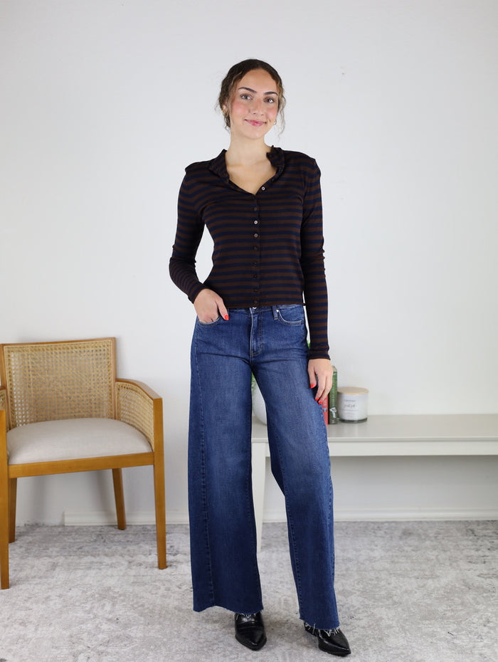A woman stands indoors on a light carpet, smiling with one hand in her pocket. She is wearing the Everette Lightweight Striped Top paired with wide-leg jeans. Behind her are a wooden chair and a white shelf adorned with books and small decor items.