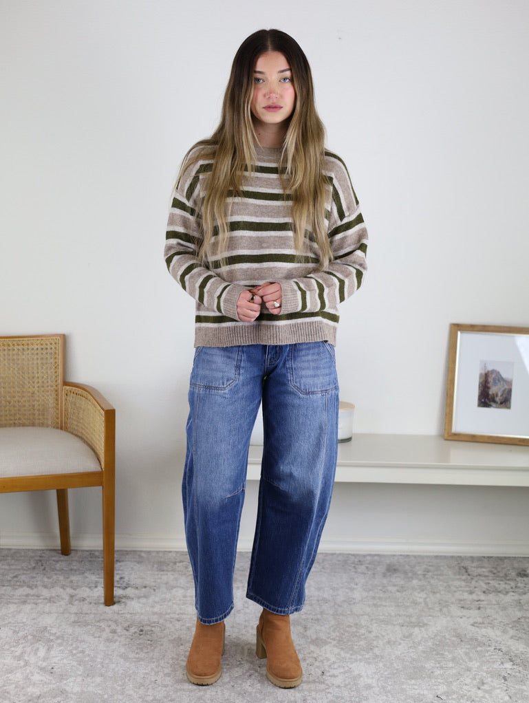 A woman with long, straight hair wears a striped sweater, Kate Double Pocket Barrel Jean, and tan boots. She stands indoors on a light rug near a wooden chair and a bench with a framed picture.