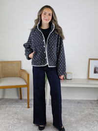 A person wears a Reba Fleece Puffer Jacket in charcoal grey with a white heart pattern, white trim, and snap buttons, paired with dark blue wide-leg jeans.