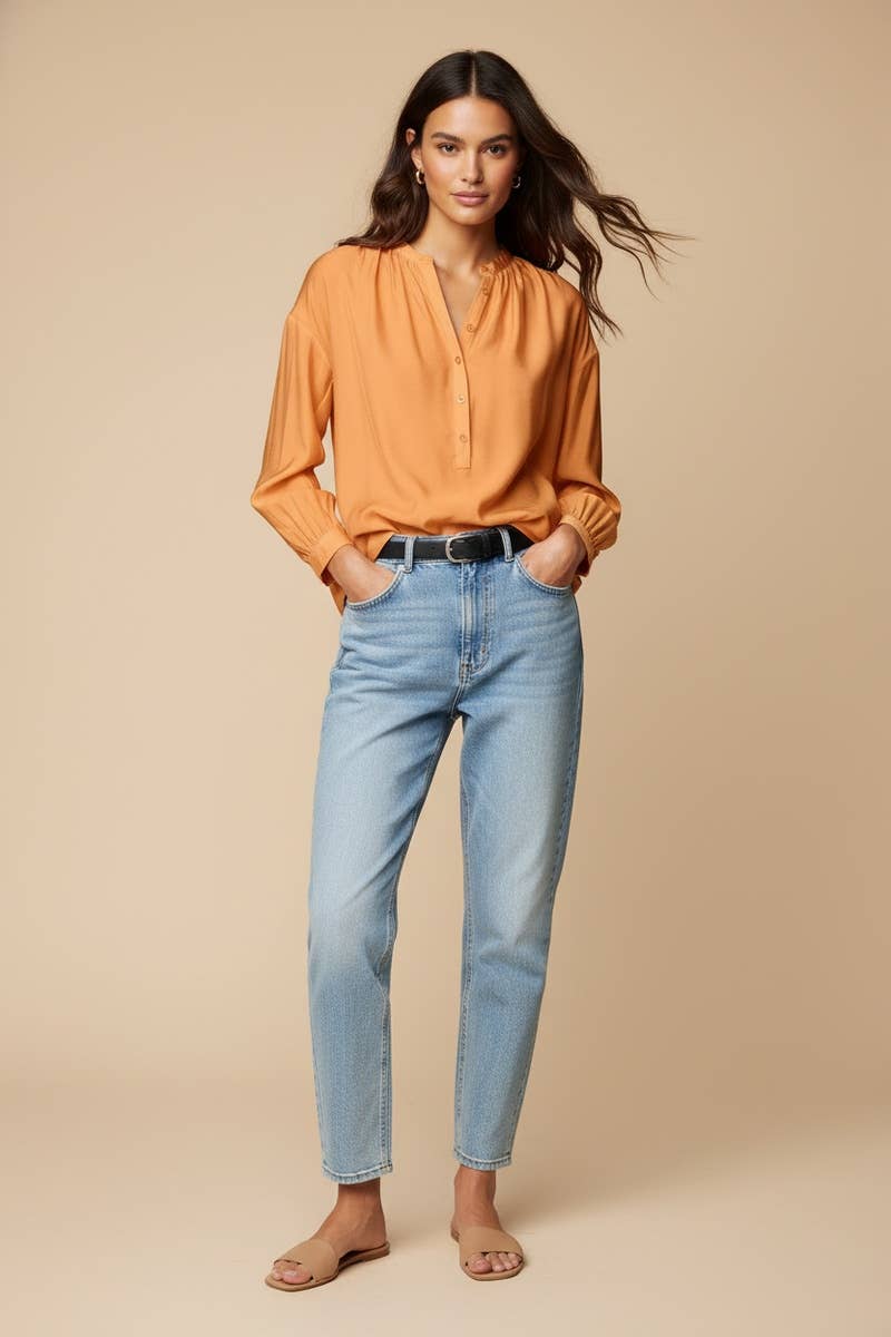 A woman wears an elegant, long-sleeved orange Naked Zebra - Button-down Blouse With Gathered Neckline tucked into light-wash denim jeans and tan sandals.