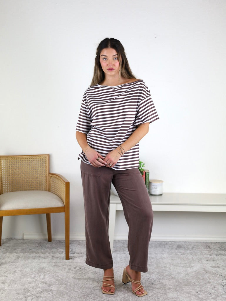 A woman stands indoors wearing the Hayden Asymmetrical Off-Shoulder Boxy Cotton Tee with stripes and loose brown pants, paired with sandals. She is standing in front of a white wall, beside a wooden chair and a white console table adorned with decorative items.
