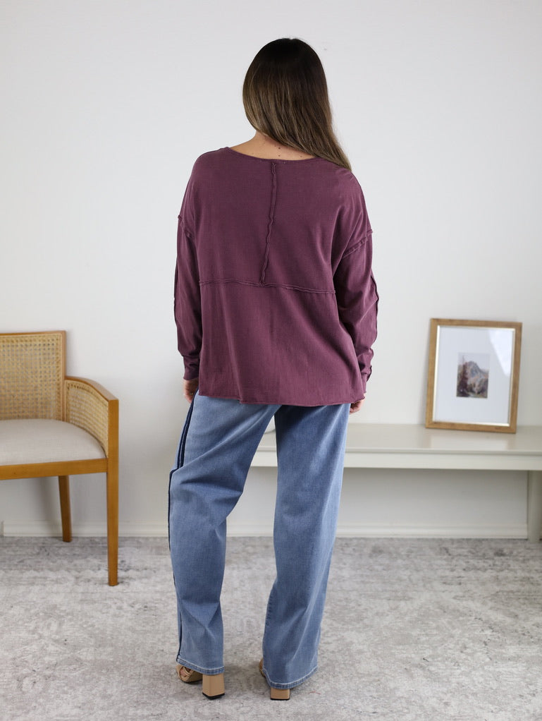 A woman faces away indoors, wearing the Ester Raw Seam Henley in maroon with light blue jeans. She stands near a wooden chair and a white shelf with a framed photo, in a room with neutral walls and carpet.