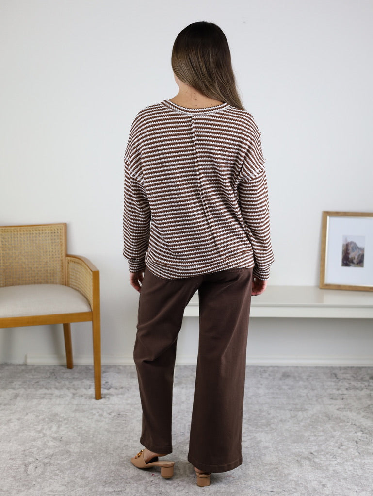 A woman with long hair stands indoors facing away, wearing the Lennon Stripe Knit Top paired with wide-leg brown pants and beige shoes. A chair, bench, and framed picture are in the background.