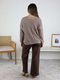 A woman with long hair stands indoors facing away, wearing the Lennon Stripe Knit Top paired with wide-leg brown pants and beige shoes. A chair, bench, and framed picture are in the background.