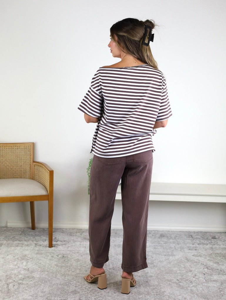 A woman stands indoors facing away, wearing the Hayden Asymmetrical Off-Shoulder Boxy Cotton Tee with black-and-white stripes, paired with brown pants, beige heels, and a hair clip. A wooden chair and a white bench are nearby on a light gray carpet.