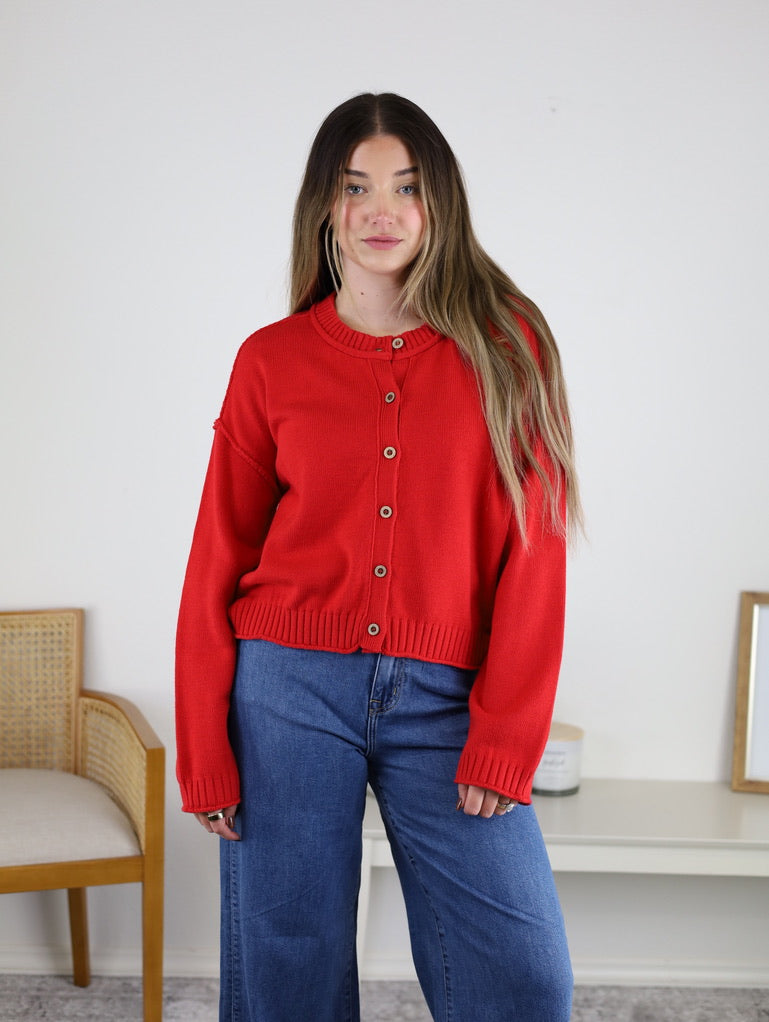 A person wearing a long-sleeved red Jude Raw Edge Cardigan and blue jeans.
