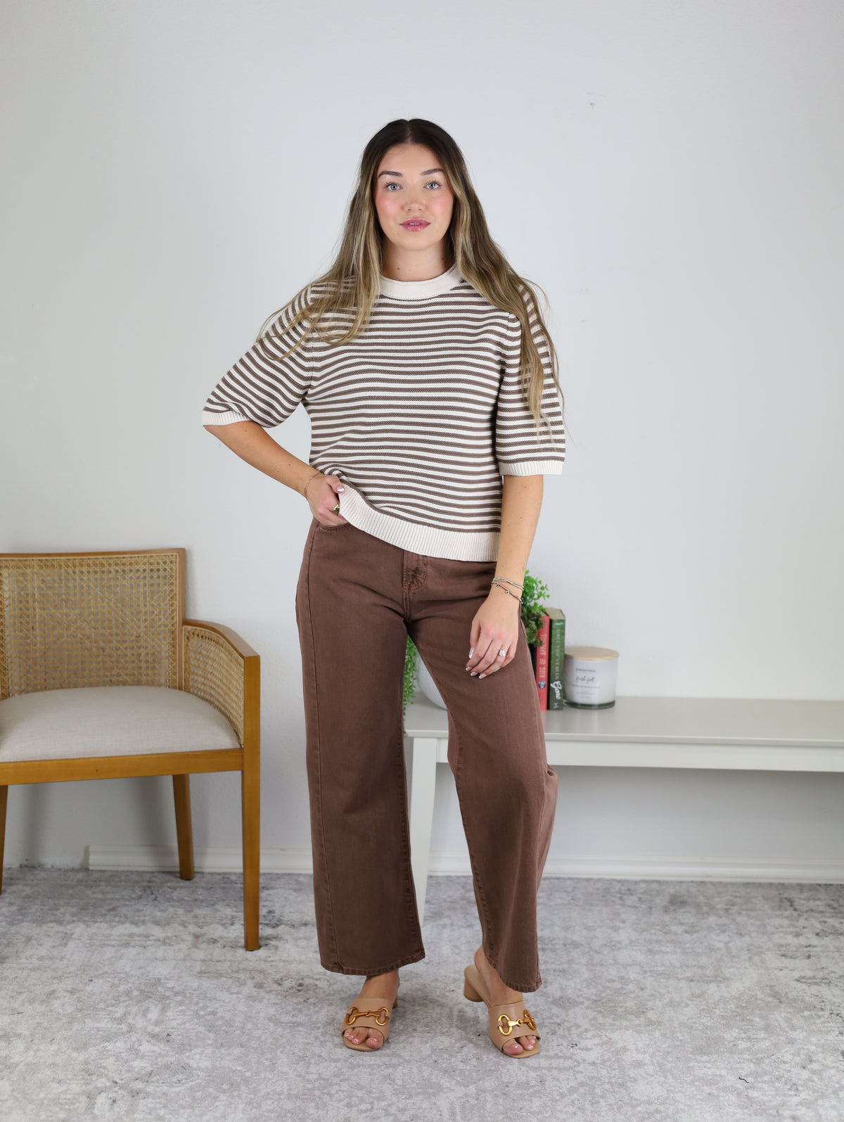 A woman stands indoors wearing the Scout Striped Sweater, featuring beige and brown cotton knit stripes, paired with brown wide-leg pants and tan sandals. She is next to a wooden chair and a white shelf adorned with plants and candles, set against a plain white wall.