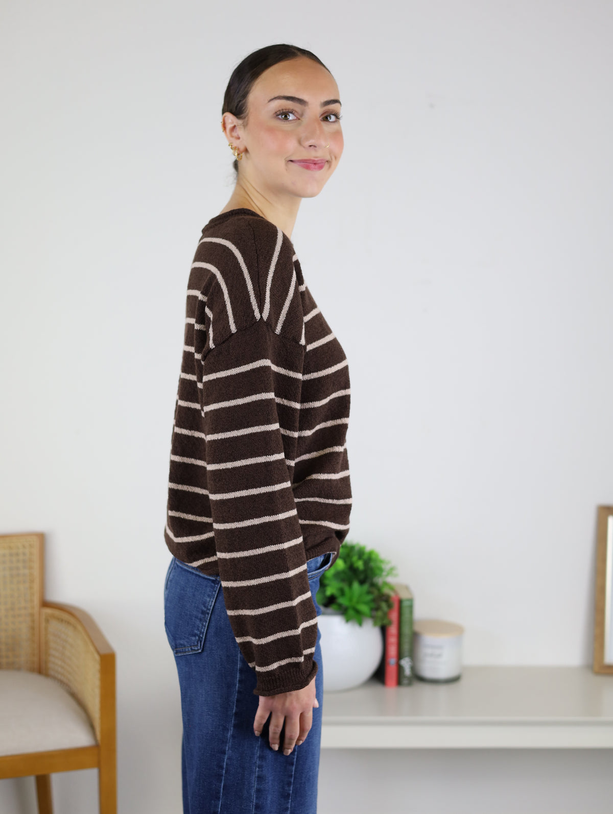 A woman with dark hair in a bun, wearing the Odette Stripe Sweater in brown and white stripes paired with blue jeans, stands sideways and smiles. Indoors, with books, a plant, and a candle on the table behind her, the cozy cotton knit of her relaxed Odette Stripe Sweater enhances the inviting atmosphere of the room.