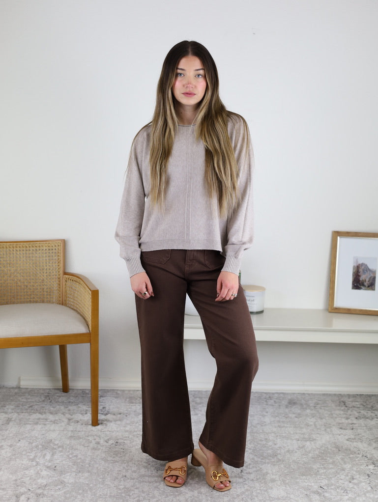 A woman wears an Eli Front Seam Sweater in beige viscose blend knit with a vertical front seam, paired with brown wide-leg pants.