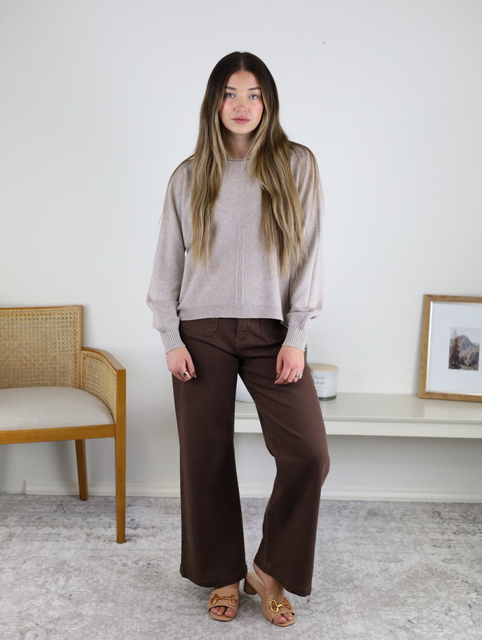 A woman wears an Eli Front Seam Sweater in beige viscose blend knit with a vertical front seam, paired with brown wide-leg pants.
