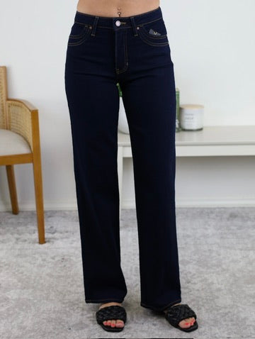 A person wearing the Mae Double Pocket Full Length Wide Leg denim jeans in dark blue with black sandals stands on a light gray carpet. In the background, there is a white table, a candle, a green plant, and part of a wooden chair.