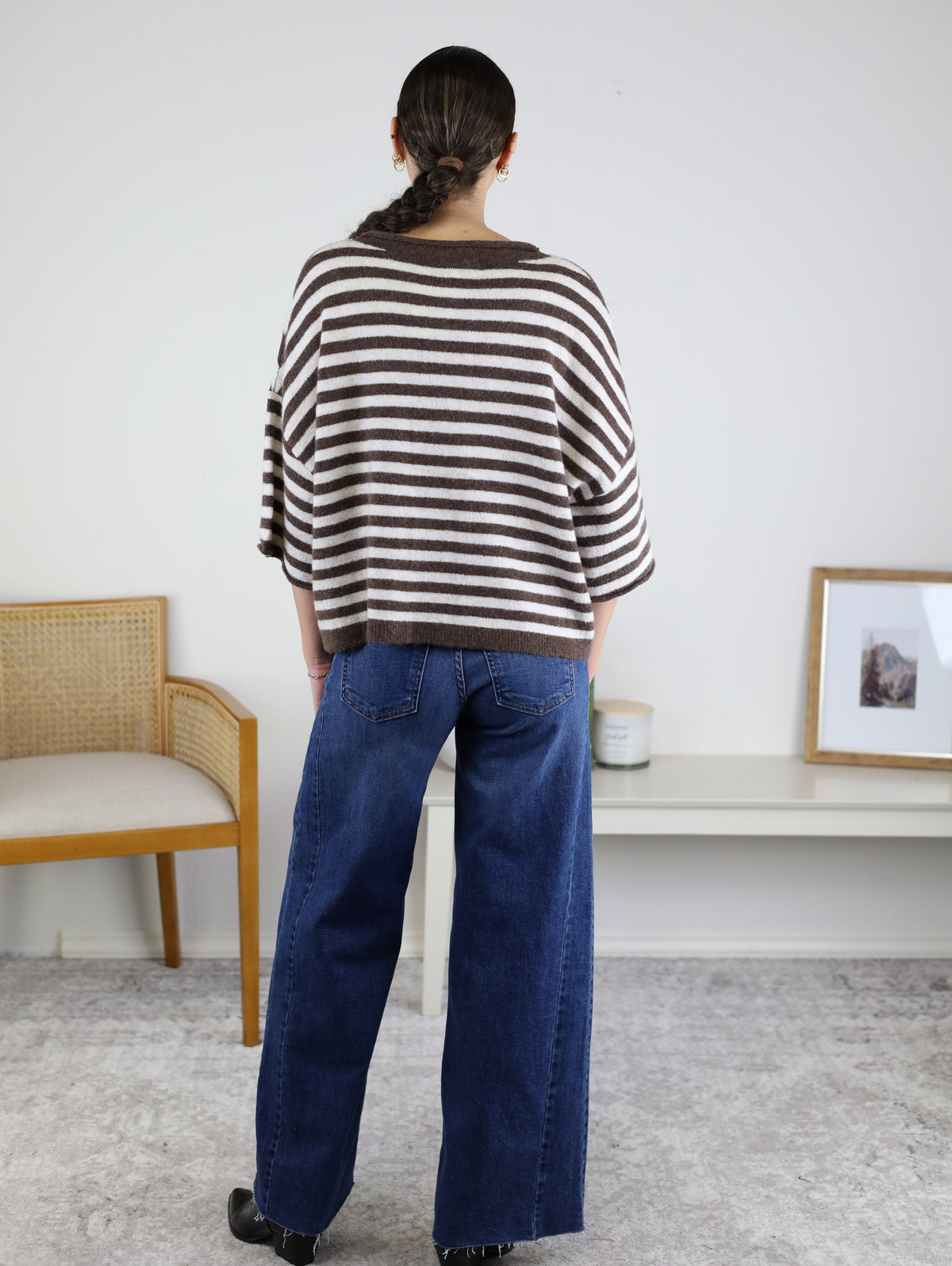 A person with a braided ponytail, wearing the Stephanie Striped Cardigan in chocolate brown stripes and blue jeans, stands facing away in a minimalist room with a chair, shelf, framed photo, and candle.