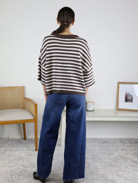 A person with a braided ponytail, wearing the Stephanie Striped Cardigan in chocolate brown stripes and blue jeans, stands facing away in a minimalist room with a chair, shelf, framed photo, and candle.