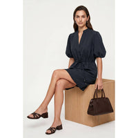 A person wears a Naked Zebra - Puff Sleeve Cinched Waist Mini Dress: Navy / L, sitting on a wooden cube while holding a brown handbag and wearing heeled sandals.