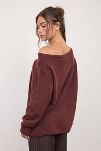 A woman with long brown hair styled in a loose bun wears The Workshop Studio’s Boat Neck Oversized Knit Pullover Sweater in Red Wood, paired with brown pinstripe pants. She looks down with a neutral expression against a plain light background.