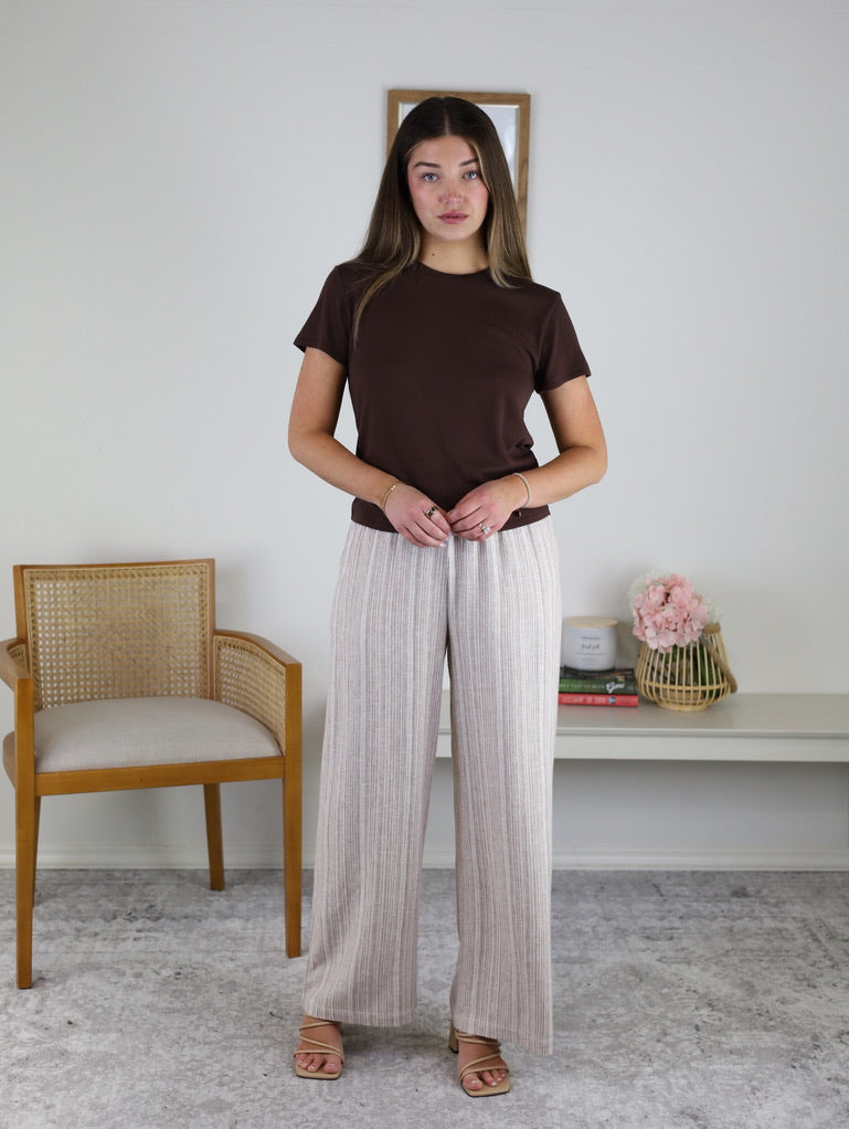A woman wears a brown short-sleeved t-shirt and Louise Linen Blend Pant, which feature thin vertical stripes and an elastic drawstring waist.