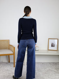 A woman with a braided ponytail stands facing away, wearing the Deepsea Ribbed Collared Knit Top—navy, fitted, and collared—paired with wide-leg jeans. She is in a minimal room featuring a chair, a bench, a framed photo, and a candle.
