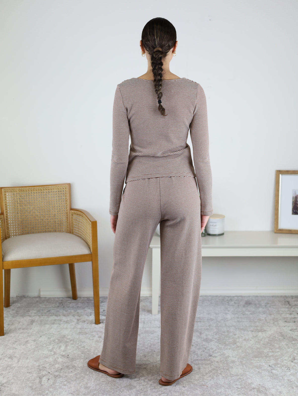 A woman with a braid stands facing away, wearing the Melissa Lounge Stripe Button Down Set in taupe—a coordinated long-sleeve top and matching pants. She stands on a light carpet near a chair and a white table with decor.