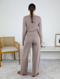 A woman with a braid stands facing away, wearing the Melissa Lounge Stripe Button Down Set in taupe—a coordinated long-sleeve top and matching pants. She stands on a light carpet near a chair and a white table with decor.