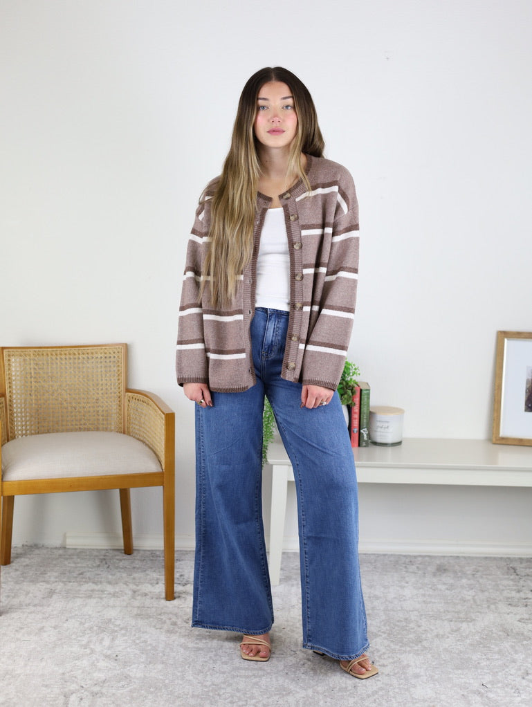A person wearing a brown and white Nora Stripe Button Down Knit Cardigan over a white tank top with wide-leg blue jeans.
