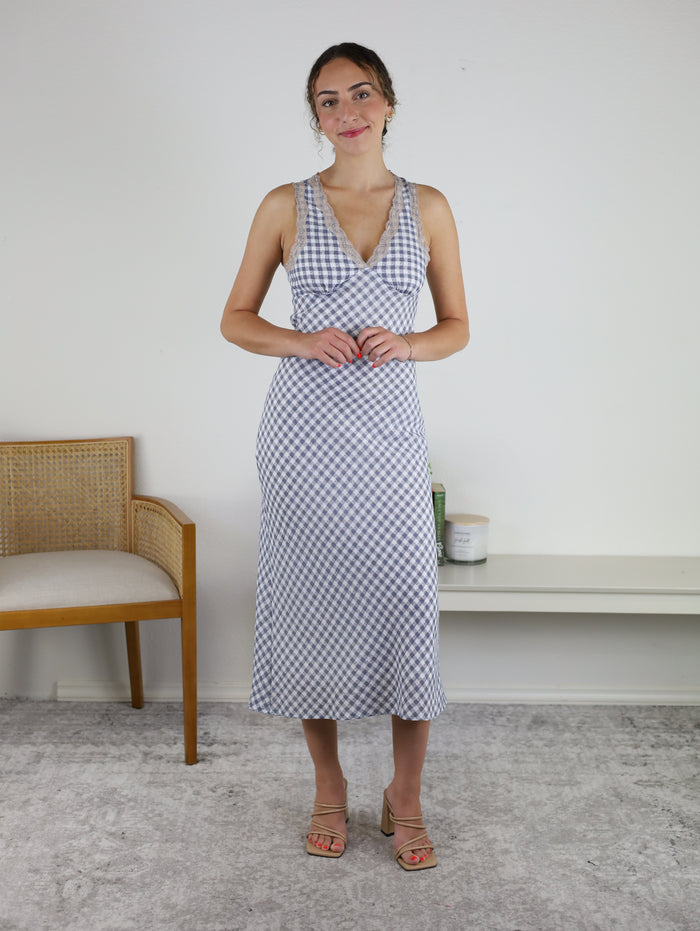 A woman stands indoors wearing the Tessa Sleeveless V-Neck Midi dress with a checkered print, paired with sandals. She is smiling slightly, with her hands clasped. A wooden chair and shelves with candles are visible in the background.