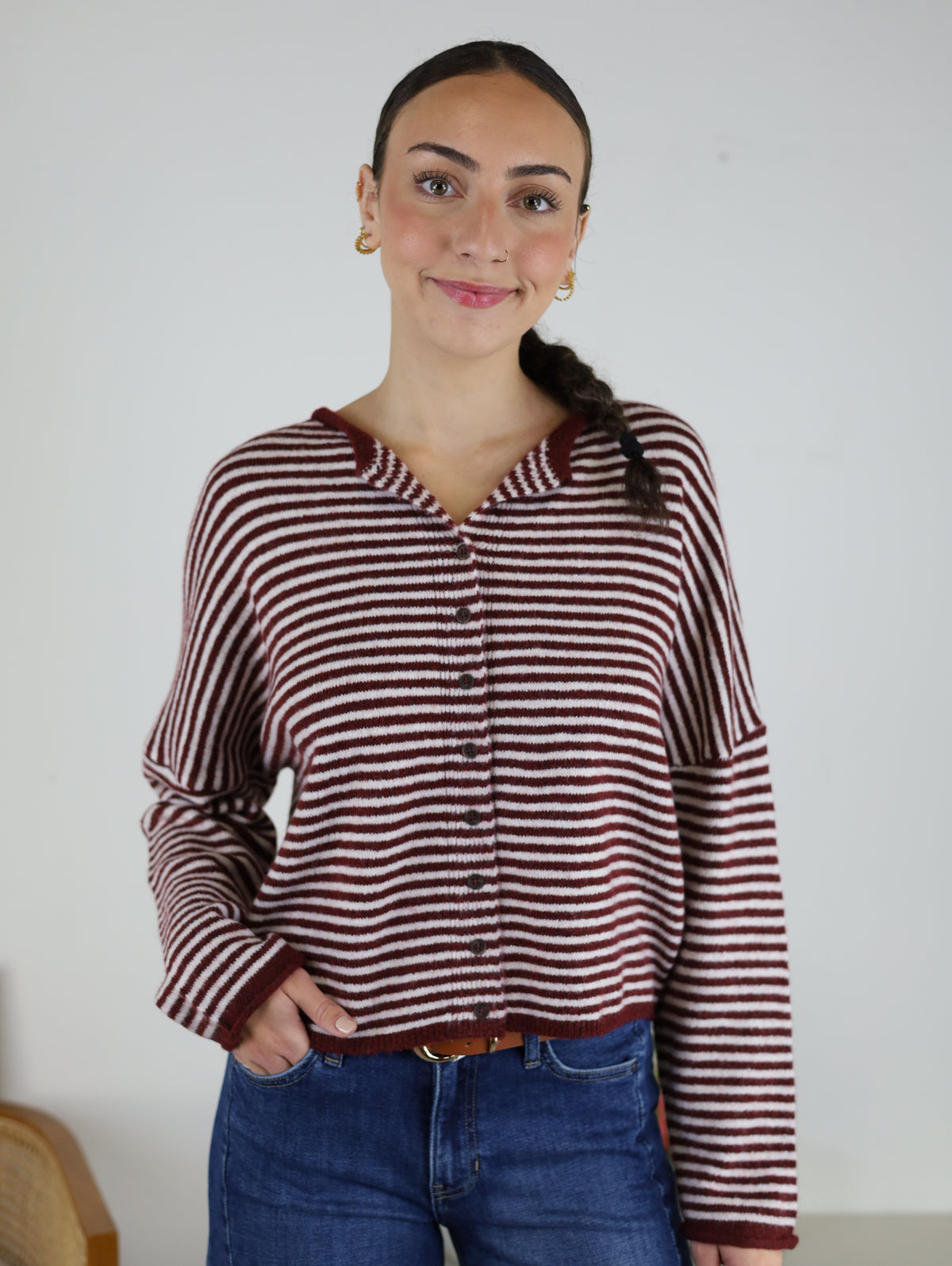 A woman with braided hair and gold hoop earrings stands smiling in front of a plain white wall, wearing blue jeans and the relaxed fit Maren Striped Cardigan—a maroon and white wool-blend knit.