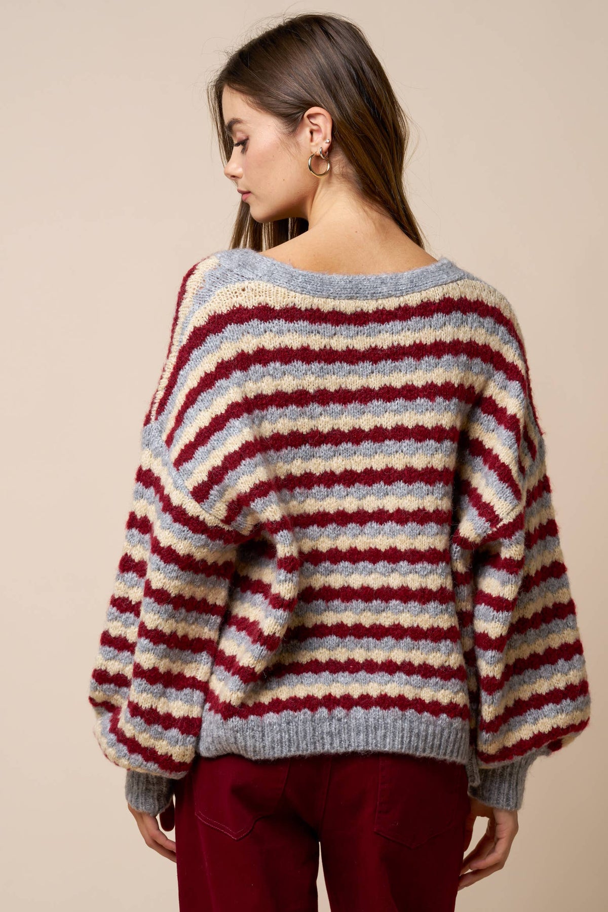 A woman with long brown hair and gold hoop earrings stands with her back to the camera against a plain beige background, wearing the Lalavon Relaxed Stripe V-Neck Button Cardigan in grey, burgundy, and beige (size L) paired with burgundy pants.